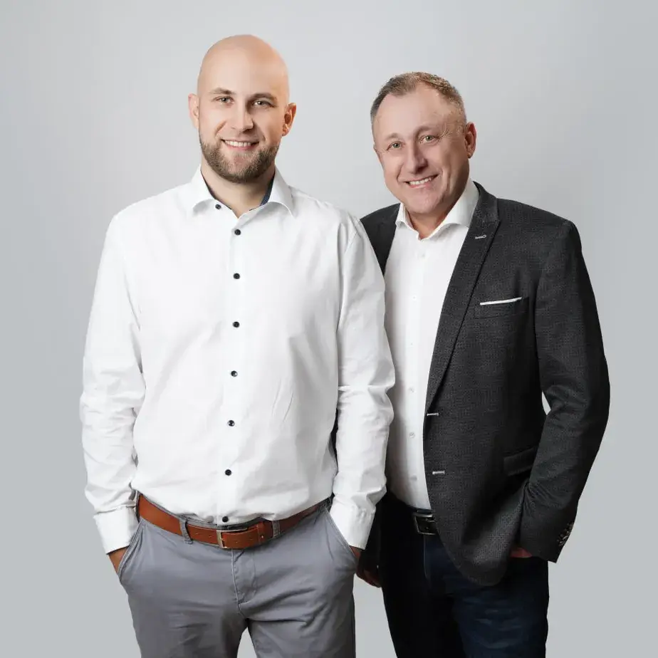 Zwei Männer stehen nebeneinander und lächeln, einer trägt weißes Hemd mit grauer Hose, der andere grauen Sakko mit Jeans